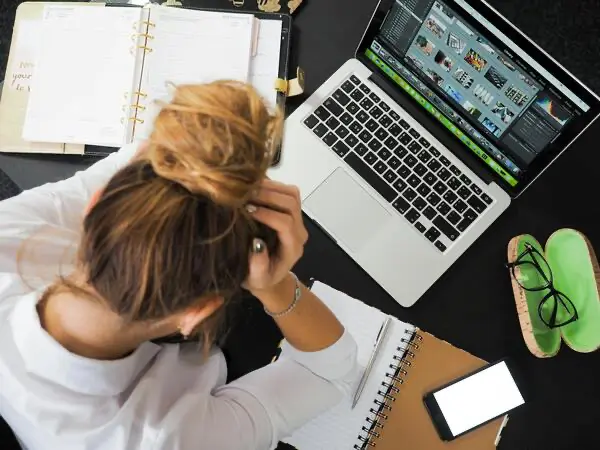 Person, die mit einem geöffneten Laptop, Notizblöcken und einem Smartphone an einem Schreibtisch sitzt, den Kopf in den Händen hält und gestresst oder frustriert wirkt, weil ihr effektive Zeitmanagement-Tools fehlen für mehr Produktivität im Homeoffice